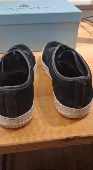 Svarta sneakers från Lanvin i mocka - Säljer ett par svarta sneakers från Lanvin med ovandel i mocka och detaljer i blankt skinn framtill. Skorna har vita platta sulor och svarta snören med metallöljetter. Snygg och stilren modell som passar till det mesta.