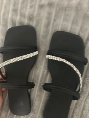 Svarta sandaler med strassdetaljer - Svarta sandaler med fyrkantig tå och två tunna remmar, varav en är täckt av glittrande strass. Platt sula och stilren design som ger en trendig vibe. Perfekta för att lyfta en outfit med lite extra bling.