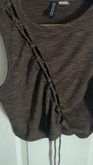 Brun ribbad croppad linnetopp H&M - Säljer en brun croppad linnetopp från H&M Divided i storlek S. Toppen är ribbad och har en cool snörningsdetalj diagonalt framtill som ger en unik look. Materialet är mjukt och stretchigt, perfekt för att styla med jeans eller kjol. Ärmlös och med rund hals.