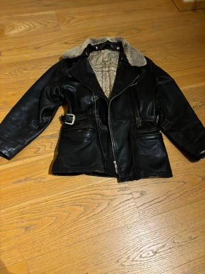 Svart skinnjacka med teddykrage - Svart skinnjacka i äkta svinläder med grym teddykrage och kartmönstrat innerfoder. Jackan har dragkedja, bältesdetaljer i midjan och flera fickor framtill. Fodret är gjort av nylon och har ett unikt kartmotiv. Perfekt för dig som gillar retrovibe och coola detaljer. Storlek S men passar för större storlekar också.