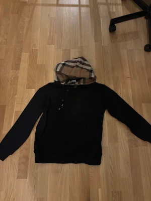 Svart burrbery hoodie med rutig huva, strl M - Svart burrbury hoodie med klassisk känguruficka och långärmad passform. Huvan har ett snyggt rutigt mönster i beige, svart, vitt och rött, samt snörning för justering. Perfekt för dig som gillar streetstyle och vill sticka ut med en unik detalj.