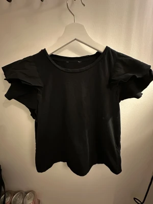 Svart blus med volangärmar - Säljer en svart blus med rund halsringning och korta volangärmar som ger en cool och trendig look. Toppen är gjord i mjuk bomull och har en loose passform. Perfekt för dig som gillar stilrena plagg med lite extra detaljer på ärmarna.
