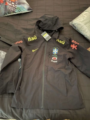 Svart Nike Brasil vindjacka med huva - Svart vindjacka från Nike med Brasil CBF-logga, sponsormärken som Itaú och Guaraná Antarctica. Jackan har huva, hel dragkedja och sportig look. Perfekt för dig som gillar fotboll.