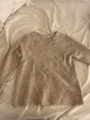 Beige ribbad stickad tröja från ONLY - Mysig beige stickad tröja från ONLY i storlek XS. Tröjan har ett ribbat mönster och v-ringad hals, samt långa ärmar. Perfekt för dig som gillar enkel och stilren design med en neutral färgton.