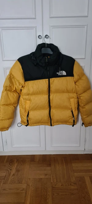 The North Face jacka  - Säljer en gul och svart pufferjacka från The North Face med hög krage och dragkedja framtill. Jackan har två sidofickor med dragkedja. Perfekt för kalla dagar och har en riktigt schysst streetstil. Den är i storlek M för barn. Så den kan passa ungdomar.