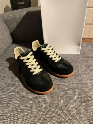 Maison Margiela svarta sneakers - Maison Margiela sneakers i svart skinn och mocka med kontrasterande vita snören och beige innersida. Snygg gummisula i brunt ger en klassisk vibe. Finns ett litet jack hör av er så skicka jag bild