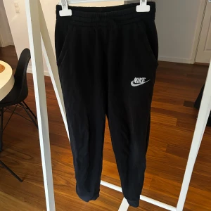 Svarta mjukisbyxor från Nike  - Svarta mjukisbyxor från Nike med broderad vit Nike-logga på benet. Byxorna har elastisk midja med snörning och sidofickor. Perfekta för chill eller träning, tillverkade i mjukt bomullsmaterial för skön komfort.