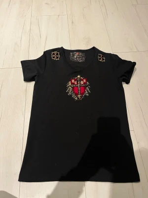Svart t-shirt med broderat korsmotiv - Svart t-shirt från KIGZ med korta ärmar och rund hals. Framsidan har ett stort broderat korsmotiv i rött, guld och svart, samt detaljer i guld och grått. På axlarna finns broderade emblem och baksidan har text i guld. Materialet känns som mjuk bomull.