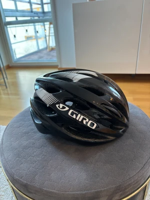 Svart cykelhjälm Giro Trinity - Säljer en svart cykelhjälm från Giro, modell Trinity. Hjälmen har en aerodynamisk form med flera ventiler för bra luftflöde och justerbara remmar. Snygga vita detaljer och logga på sidan. Perfekt för dig som vill ha både stil och säkerhet på cykelturen.