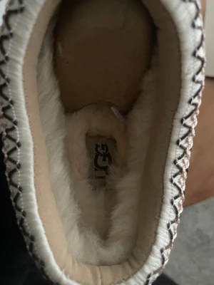 UGG beige slip-on mocka skor - Säljer ett par beige slip-on skor från UGG med mjuk päls på insidan och mocka på utsidan. Skorna har en rund tå, platt sula och dekorativt broderi runt öppningen i vitt och brunt. Perfekta för kalla dagar när du vill vara både chill och varm.