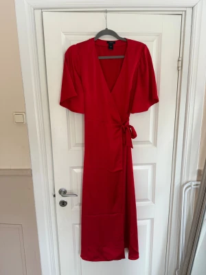 Röd omlottklänning från Lindex - Snygg röd omlottklänning från Lindex med knyt i midjan och v-ringad hals. Klänningen har korta, vida ärmar och är tillverkad i ett mjukt, lätt material som ger ett fint fall. Perfekt för dig som gillar en enkel men stilren look.