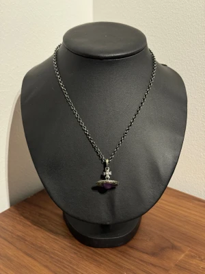 Vivienne Westwood halsband med lila orb - Snyggt halsband från Vivienne Westwood med klassisk orb-berlock i silverfärgad metall och en lila detalj i mitten. Kedjan är grov och har märkets logga vid låset. Kommer med originalförpackning, tygpåse och putsduk. Perfekt statement-piece för dig som gillar ikoniska accessoarer.