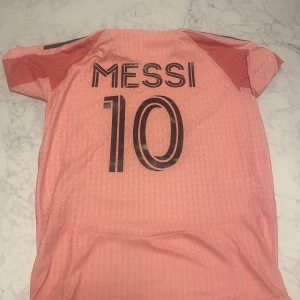 Inter Miami Messi #10 fotbollströja - Säljer en rosa Inter Miami fotbollströja från Adidas med Messi och nummer 10 på ryggen. Tröjan har svarta detaljer, klubbmärke på bröstet och klassiska Adidas-ränder på axlarna. Tillverkad i lätt och ventilerande material, perfekt för match eller träning. Oanvänd helt ny