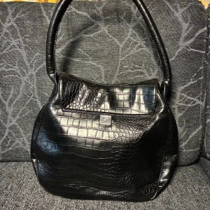 Svart axelväska i croco-mönstrat skinn - Cool svart axelväska från Sisley i croco-mönstrat skinn. Väskan har rundad form, bred axelrem och snygg metallogo framtill. Stängs med lock och knapp, perfekt för dig som vill ha en unik och stilren accessoar.