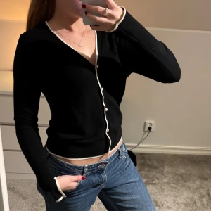 Svart kofta med vita detaljer - Snygg svart kofta med vita kanter och pärlknappar. V-ringad och har långa ärmar. Perfekt att styla med jeans eller kjol för en clean look.