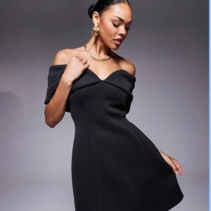 Svart offshoulder klänning från ASOS - Helt oanvänd supersnygg svart kortklänning från ASOS med offshoulder och v-ringad urringning. Klänningen har en figurnära passform upptill och lätt utsvängd kjol. Tillverkad i ett slätt material som ger en elegant look. Perfekt för dig som vill sticka ut med stilren vibe. Har även en helt oanvänd i storlek 38. Ordinariepris: 539kr 