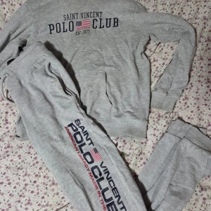 Grå mjukisbyxor Saint Vincent Polo Club - Grå mjukisbyxor från Saint Vincent Polo Club med tryck längs benet och bakficka. Byxorna har snörning i midjan, ribbade muddar vid benslut och är gjorda i mjukt bomullsmaterial. Perfekta för chill eller sportiga dagar.