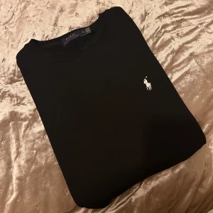 Svart sweatshirt från Polo Ralph Lauren - Svart sweatshirt från Polo Ralph Lauren i storlek 2XL men passar L. Tröjan har rund hals, lång ärm och en vit broderad logga på bröstet. Tillverkad i mjuk bomull som är skön mot huden. Enkel och stilren design som funkar till många olika looks.