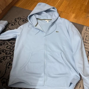 Ljusblå hoodie från Lacoste M - Säljer en ljusblå hoodie från Lacoste i klassisk passform. Tröjan har huva, dragkedja framtill och två stora fickor. Materialet är mjuk bomull och den har det ikoniska Lacoste-krokodilemblemet på bröstet. Perfekt för chill dagar och enkel att matcha.