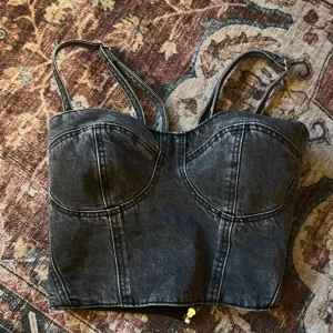 Svart denim korsett från NA-KD - Säljer en svart korsett-topp från NA-KD i denim med markerade sömmar och justerbara axelband. Toppen har en snygg bustierform och dragkedja i sidan för enkel påklädning. Perfekt för dig som vill ha en edgy och trendig look.
