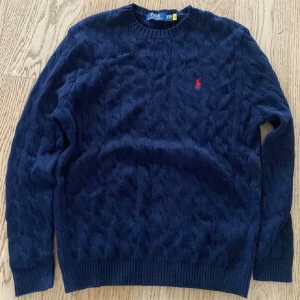 Marinblå stickad tröja från Polo Ralph Lauren - Säljer en marinblå stickad tröja från Polo Ralph Lauren med klassiskt kabelstickat mönster och röd broderad logga på bröstet. Tröjan har rund hals, långa ärmar och ribbade muddar vid ärmslut och nederkant. Perfekt för dig som gillar stilren och tidlös design.❌r3epl1c@ ❌