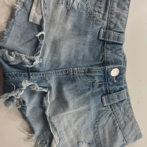 Ljusblå jeansshorts från Gina Tricot - Säljer ett par ljusblå jeansshorts från Gina Tricot i storlek XXS. De har råa, fransiga kanter och slitna detaljer för en cool look. Klassisk femficksmodell med bälteshällor och knappstängning. Perfekta för varma dagar!