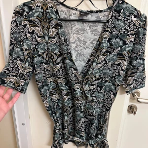 Blommig blus från Gina Tricot Strl: S - Supersöt blus från Gina Tricot med blommigt mönster i svart, grönt och beige. Blusen har v-ringning, korta ärmar och markerad midja. Perfekt för dig som gillar unika prints och vill sticka ut lite extra.