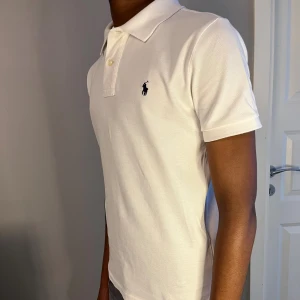 Ralph Lauren piké slim fit - vit  - Klassisk Ralph Lauren piké i vit färg med svart broderad logga på bröstet. Storlek: 14/16 år (motsvarar ungefär XS) | Skick: Nyskick – använd 1–2 gånger, inga fläckar eller slitage | Material: Bomull | Passar både unisex och ger en clean, tidlös look.