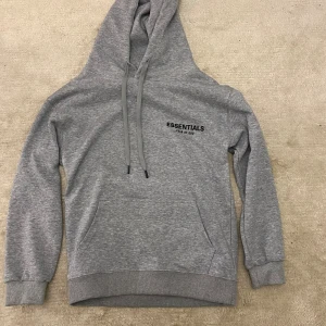 Grå Essentials Fear of God hoodie - Snygg grå hoodie från Essentials Fear of God med svart logga på bröstet och stor text på ryggen. Tröjan har huva med snörning, känguruficka och ribbade muddar. Perfekt för en chill och trendig look.