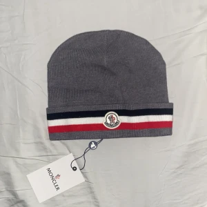 Grå Moncler mössa med ränder - Snygg grå mössa från Moncler med klassiska ränder i svart, vitt och rött längs kanten. Mössan har Moncler-logga framtill och är ribbstickad för en skön passform. Perfekt accessoar för att lyfta din vinterstil.