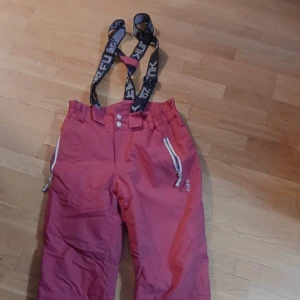 Rosa täckbyxor med hängslen - Säljer ett par rosa täckbyxor från Funk med svarta och vita justerbara hängslen. Byxorna har vita dragkedjor på fickorna och två knappar i midjan. Perfekta för vintern och snöaktiviteter. Materialet är slitstarkt syntet och designen är loose fit. Skriv jämna om ni vill ha bild med på, kom jämna med förslag:)