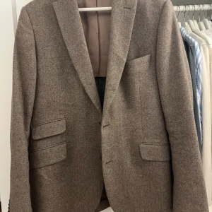 Brun kavaj från Morris Stockholm - Snygg brun kavaj från Morris Stockholm med klassisk passform och två framfickor med lock. Insidan har ett blommigt foder och broderad logga. Materialet är ull och fodret är i sidenliknande tyg. Perfekt för dig som gillar stilrena detaljer.