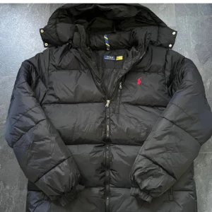 Svart dunjacka från Polo Ralph Lauren - Svart dunjacka från Polo Ralph Lauren med röd broderad logga på bröstet. Jackan har huva, tvåvägsdragkedja med RL-detalj och puffad design. Perfekt för kalla dagar och har en clean, stilren look som funkar till streetwear.