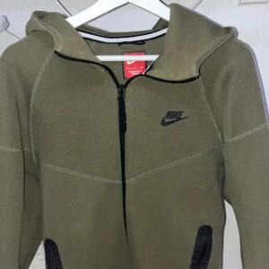 Olivgrön Nike hoodie med dragkedja - Nike hoodie i olivgrönt med hel dragkedja och svarta detaljer. Tröjan har huva, långa ärmar och två fickor med svarta dragkedjor. Perfekt för chill eller träning, med klassisk Nike-logga på bröstet.