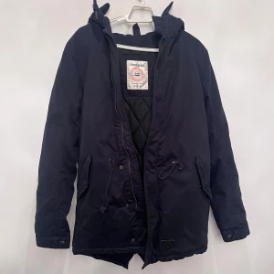 Svart parkajacka från Crocker - Svart parkajacka från Crocker med huva, snörning i midjan och quiltat foder. Jackan har två stora fickor framtill, knappdetaljer vid ärmslut och dragkedja med knappslå. Perfekt för kalla dagar med sin långa passform och stilrena look.