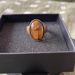 Oval ring med tigeröga-sten - Cool ring med stor oval tigeröga-sten i bruna och gyllene toner. Ringen har en bred metallram i antik guldnyans som ger en vintagekänsla. Perfekt statement piece för dig som gillar unika accessoarer med natursten.