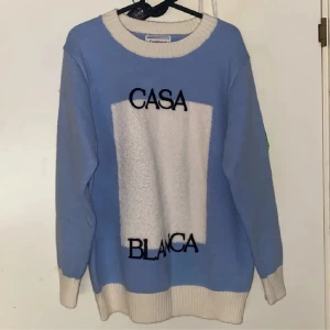 Blå stickad tröja från Casablanca - Snygg blå stickad tröja från Casablanca med kontrasterande vita ribbade muddar och krage. Framsidan har en vit rektangel med svart broderad text 'CASA BLANCA'. Tröjan har långärmad passform och känns både chill och stilren.