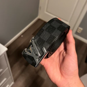 Svart rutigt Louis Vuitton bälte - Snyggt svart bälte från Louis Vuitton med klassiskt rutigt mönster och en stor LV-logga i silvermetall som spänne. Bältet är gjort i skinn och har en stilren och exklusiv känsla som lyfter vilken outfit som helst.