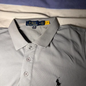 Grå pikétröja från Polo Ralph Lauren - Snygg grå pikétröja från Polo Ralph Lauren med klassisk krage och tre knappar framtill. Tröjan har korta ärmar och den ikoniska broderade loggan på bröstet. Tillverkad i 100% bomull för en skön och luftig känsla.