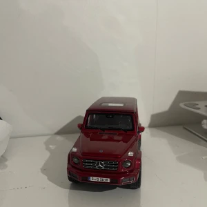 Mercedes-Benz G-Klass modellbil - Detaljerad röd modellbil av Mercedes-Benz G-Klass med V8-märkning, soltak och autentiska detaljer som registreringsskylt och reservhjul. Skicket är mycket bra. Perfekt för samlare eller som dekor.