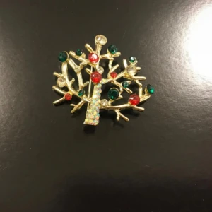 Glittrig brosch i form av träd - Unik brosch i form av ett träd med glittriga stenar i rött, grönt och vitt på en gyllene stomme. Stammen är täckt av små pärlor som ger extra bling. Perfekt för dig som vill pimpa upp en enkel outfit inför julen!