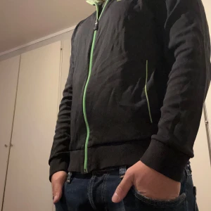 Hugo boss tröja  - Svart jacka från BOSS med grön dragkedja och gröna detaljer vid fickorna. Jackan har hög krage, lång ärm och BOSS-logga broderad på bröstet. Perfekt för dig som gillar stilrena plagg med en twist.