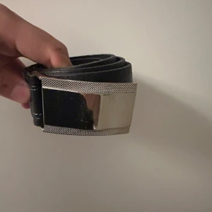 Svart bälte med silverspänne - Snyggt svart bälte med ett brett, blankt silverspänne som har en cool textur runt kanterna. Bältet är stilrent och passar perfekt till jeans eller andra byxor. Materialet är skinn och spännet är i metall med en modern känsla.