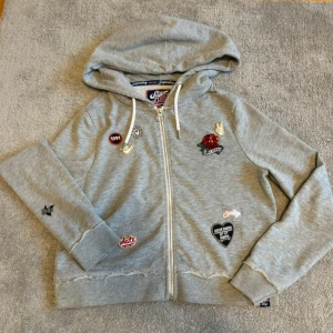 Unik Grå hoodie med stickers från Superdry - Grå hoodie från Superdry med coola broderade patches och dragkedja framtill. Tröjan har huva med snörning och ribbade muddar. Materialet är mjuk bomull och polyester, perfekt för chill dagar. Snygga detaljer som ger streetkänsla.
