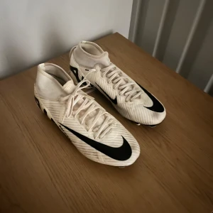 Nike Mercurial Air Zoom fotbollsskor - Säljer ett par Nike Mercurial Air Zoom fotbollsskor i ljusbeige med svarta detaljer och stor Swoosh-logga. Skorna har snörning, platt sula med dobbar och en strumpliknande krage för extra stöd. Tillverkade i syntetmaterial för lätt känsla och bra grepp på planen.
