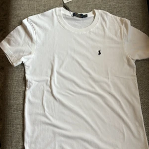 Vit t-shirt från Polo Ralph Lauren - Vit t-shirt från Polo Ralph Lauren med klassisk rund hals och korta ärmar. T-shirten är gjord i mjuk bomull och har den ikoniska broderade loggan i svart på bröstet. Enkel och stilren design som funkar till allt.