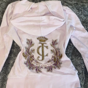 Ljusrosa Juicy Couture hoodie i velour - Supermjuk ljusrosa hoodie från Juicy Couture i velour med ikoniskt broderi och strass på ryggen. Guldig dragkedja med J-hänge, huva och kängurufickor framtill. Perfekt för dig som älskar den klassiska Y2K-looken!