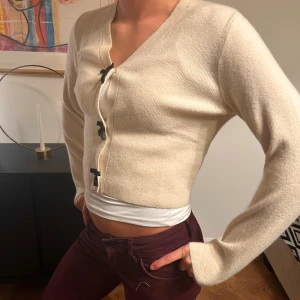 Beige kofta med rosettknäppning - Superfin beige kofta med croppad passform och långa ärmar. Koftan har v-ringning och stängs med tre svarta rosetter framtill. Mjuk och stickad känsla, perfekt för lager-på-lager. Enkel att styla till både jeans och kjol.