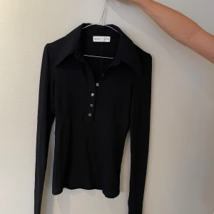 Svart långärmad polotopp från H&M Studio - Snygg svart polotopp från H&M Studio med lång ärm och klassisk krage. Toppen har en ribbad struktur och knappar framtill som ger en cool retrovibe. Perfekt att styla med jeans eller kjol för en trendig look.