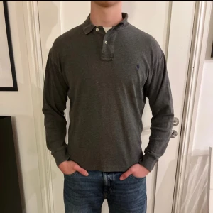 Grå pikétröja från Polo Ralph Lauren - Snygg grå långärmad pikétröja från Polo Ralph Lauren med klassisk krage och knäppning framtill. Tröjan har den ikoniska broderade loggan på bröstet och är gjord i mjuk bomull. Perfekt för en clean och stilren look.